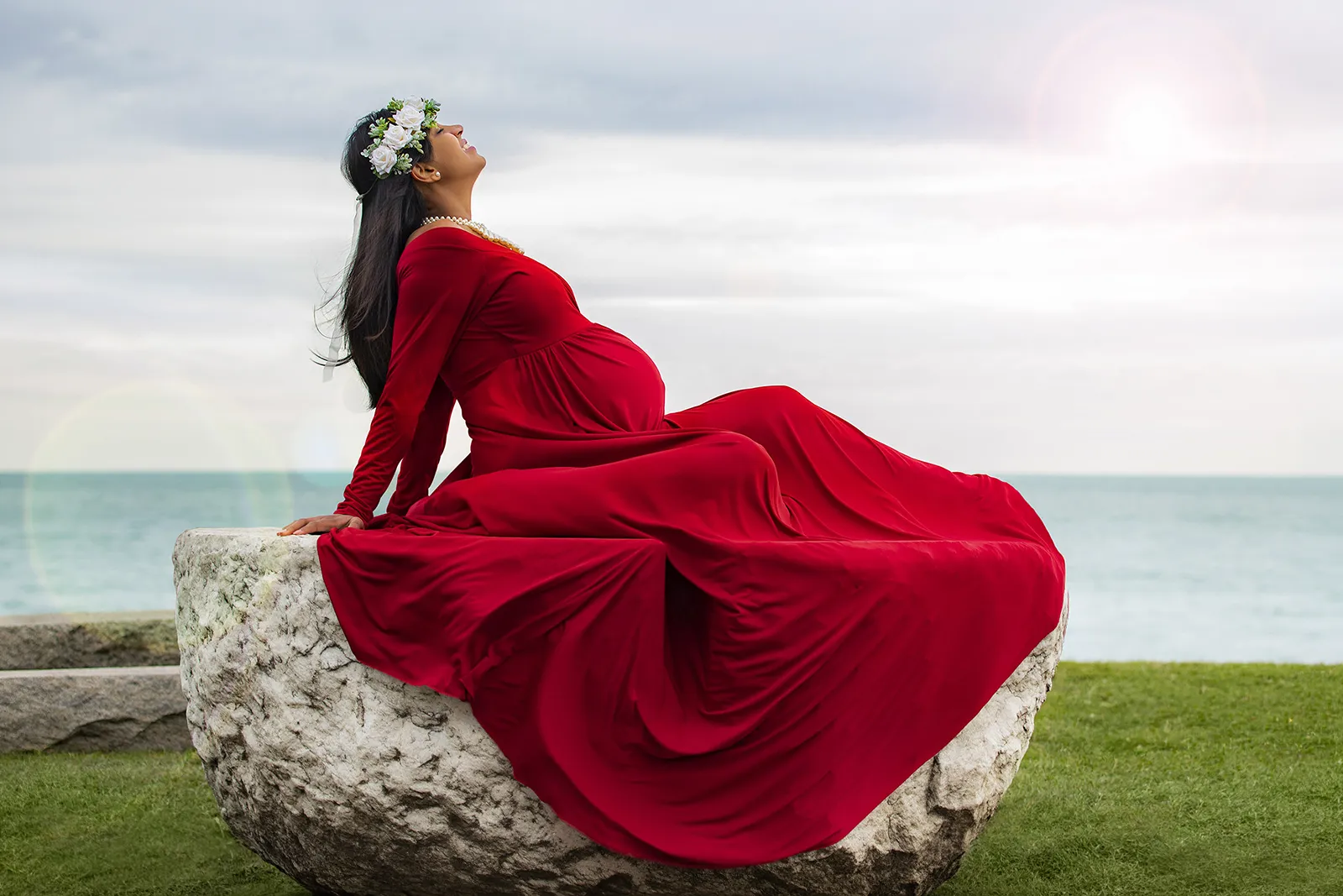 Profile view of a pregnant mother sitting on top of a rock wearing a red gown enjoying the moment smiling in Glencoe