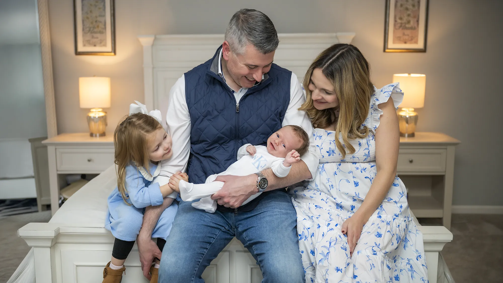 family of four sitting at the edge of a bed. father is holding a newborn baby boy smiling. wife and toddler daughter are smiling sitting next to the father. they're all looking at the baby
